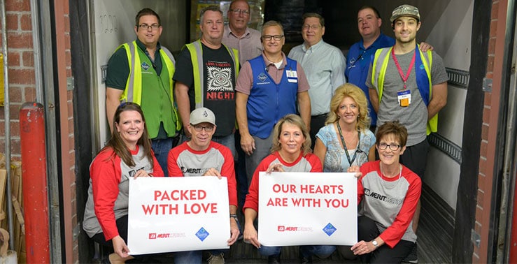 A group of Merit employees shows signs that read "Packed with Love" and "Our Hearts are With You"