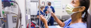 Masked nurse setting up medical devices, with two other individuals in the background