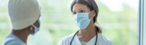 Women - one a masked physician in white, the other a cancer patient with a mask and a yellow cap on - talking in front of a window
