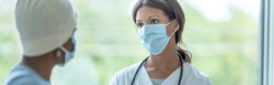 Women - one a masked physician in white, the other a cancer patient with a mask and a yellow cap on - talking in front of a window